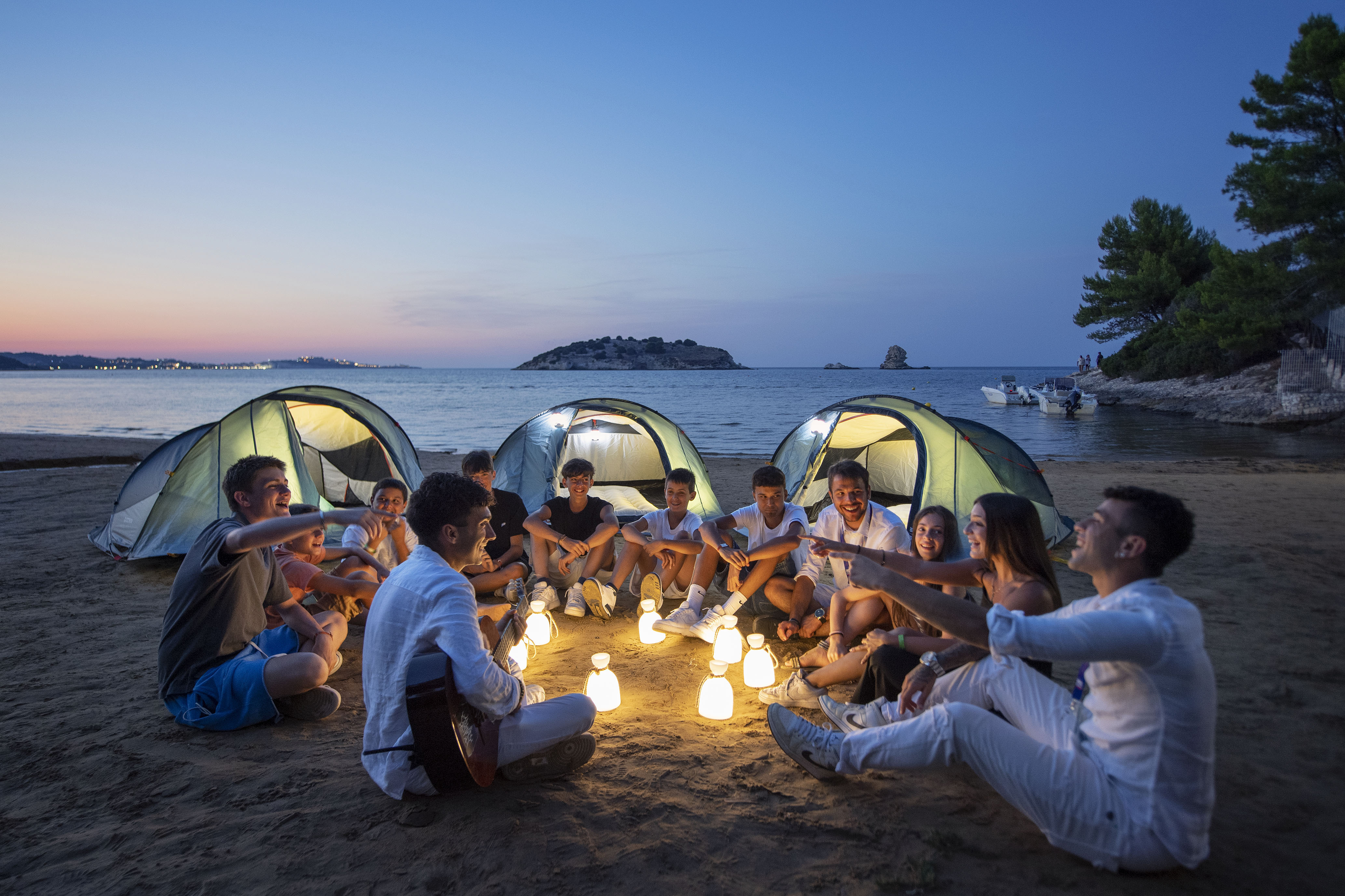 Giovani sulla spiaggia al tramonto, seduti in cerchio intorno a lanterne con chitarra e tende.